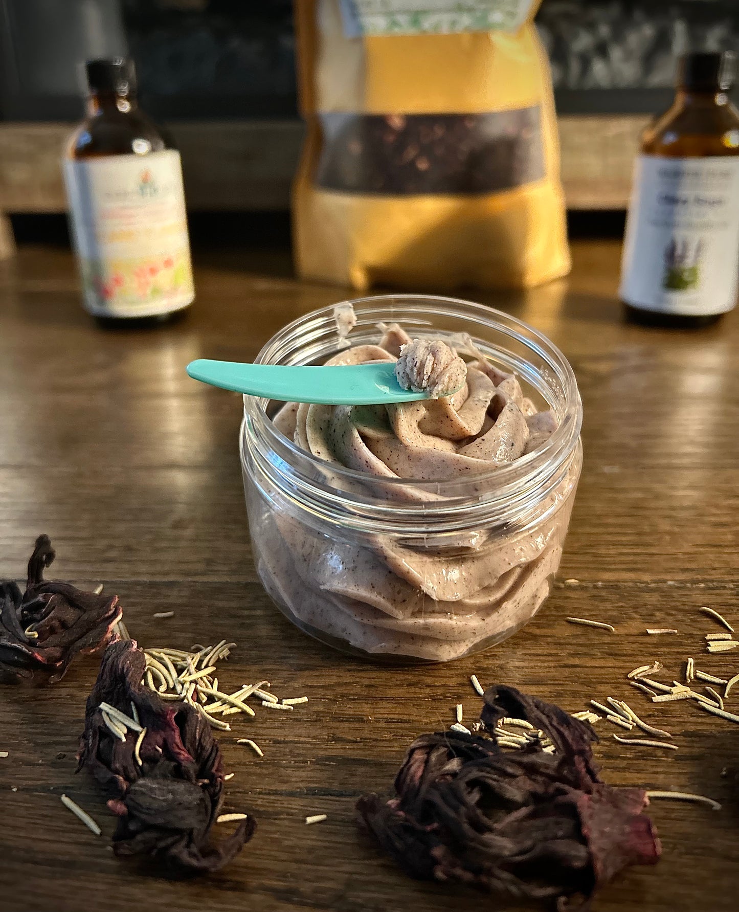 Whipped body butter in a jar with a teal spoon, rose petals, and bottles blurred in the background.