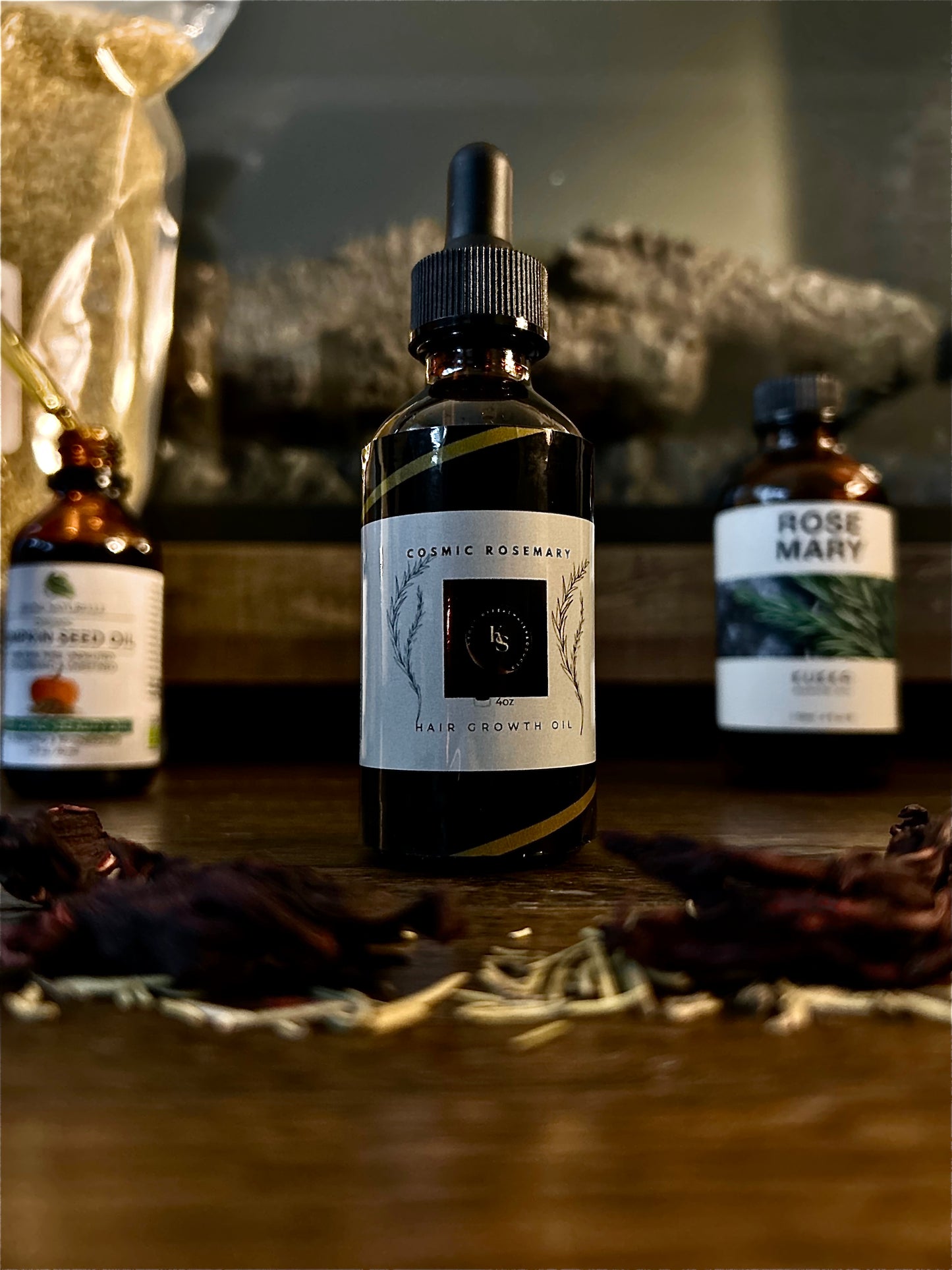 A dropper bottle labeled "Cosmic Rosemary Hair Growth Oil" prominently displayed with other bottles and herbs in soft focus.